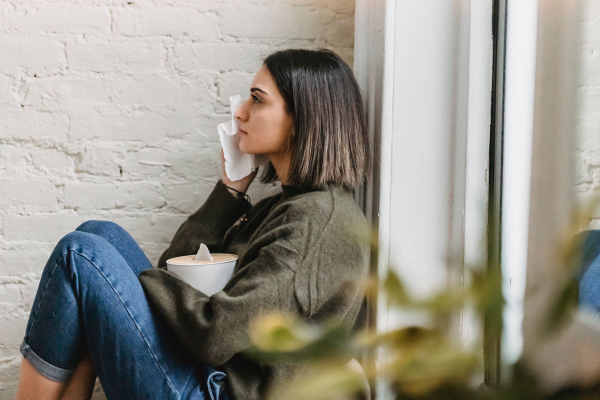 Side view of depressed young ethnic female millennial in stylish clothes wiping tears away with tissue while sitting alone near window at home
