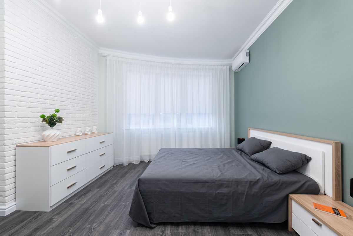 Contemporary bedroom interior with bed against table and chest of drawers on parquet under shiny light bulbs