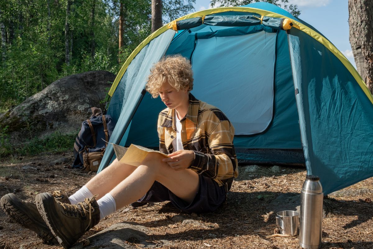 Teen sitting by tent in forest, reading a map during a summer camping trip.