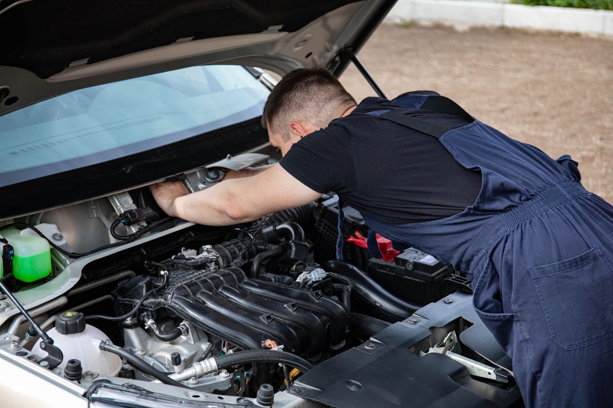 A mechanic in overalls works on a car engine outdoors, showcasing automotive maintenance.