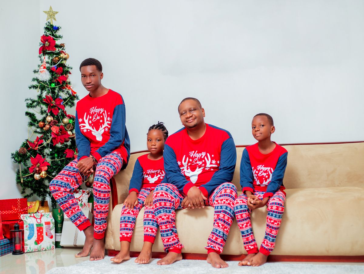 Family of four in matching Christmas pajamas sitting by a decorated tree indoors.