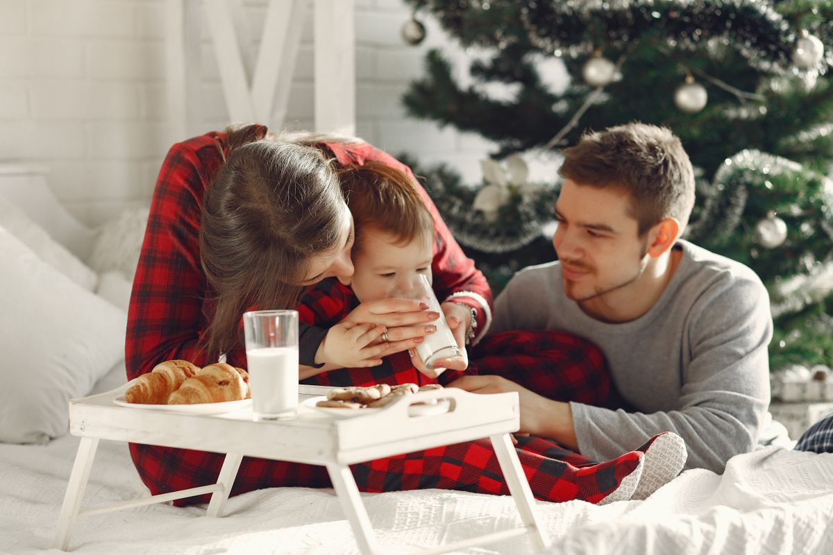A family shares a cozy Christmas morning in bed with snacks by the festive tree.