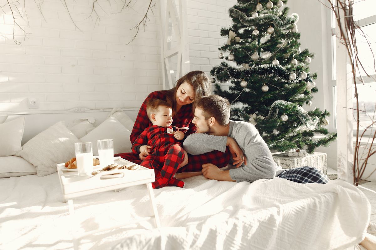 Family enjoying Christmas morning together, sitting on bed in festive pajamas near decorated tree.
