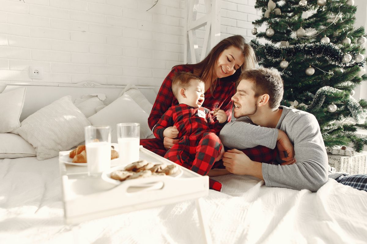 Family enjoying breakfast in bed with holiday spirit, next to a decorated Christmas tree.