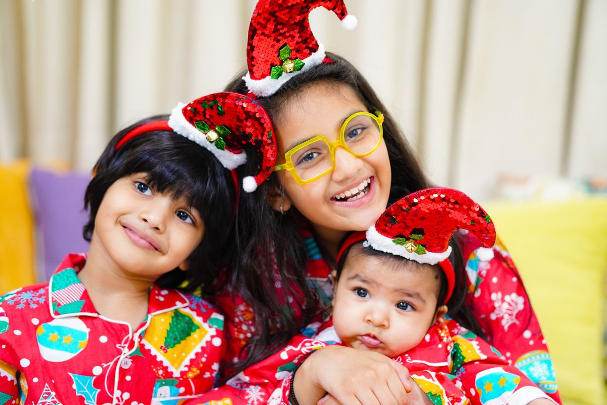 Three smiling siblings in matching Christmas pajamas and festive Santa hats, embracing the holiday spirit indoors.