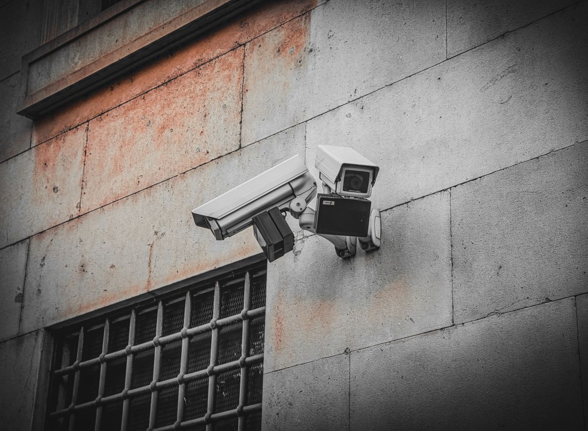 Two surveillance cameras mounted on an aged concrete wall beside a barred window, capturing urban security.