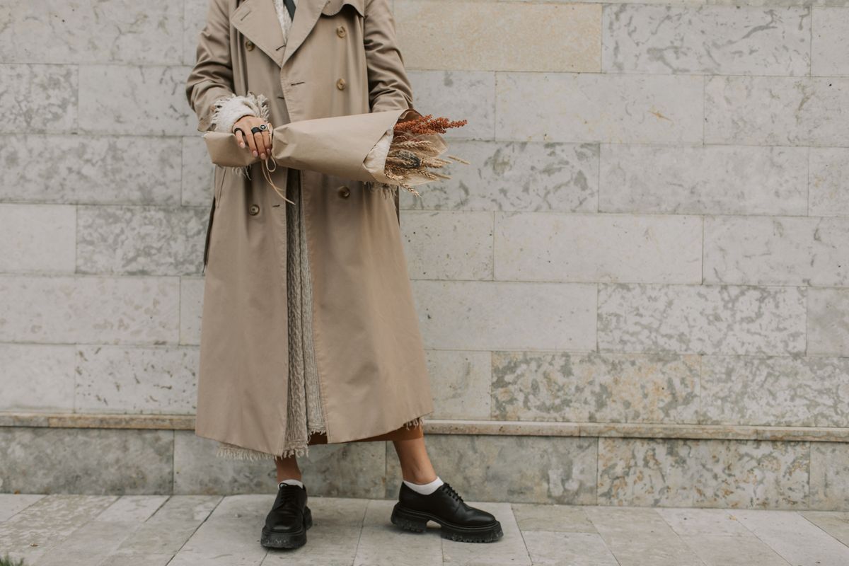Fashionable woman in a trench coat holding dried flowers against a marble wall.