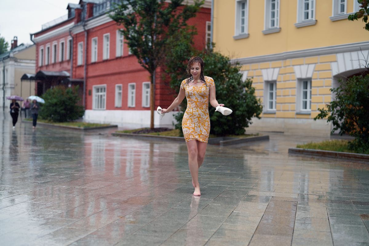 Smiling woman in yellow dress walks barefoot in a rainy urban street, holding shoes.