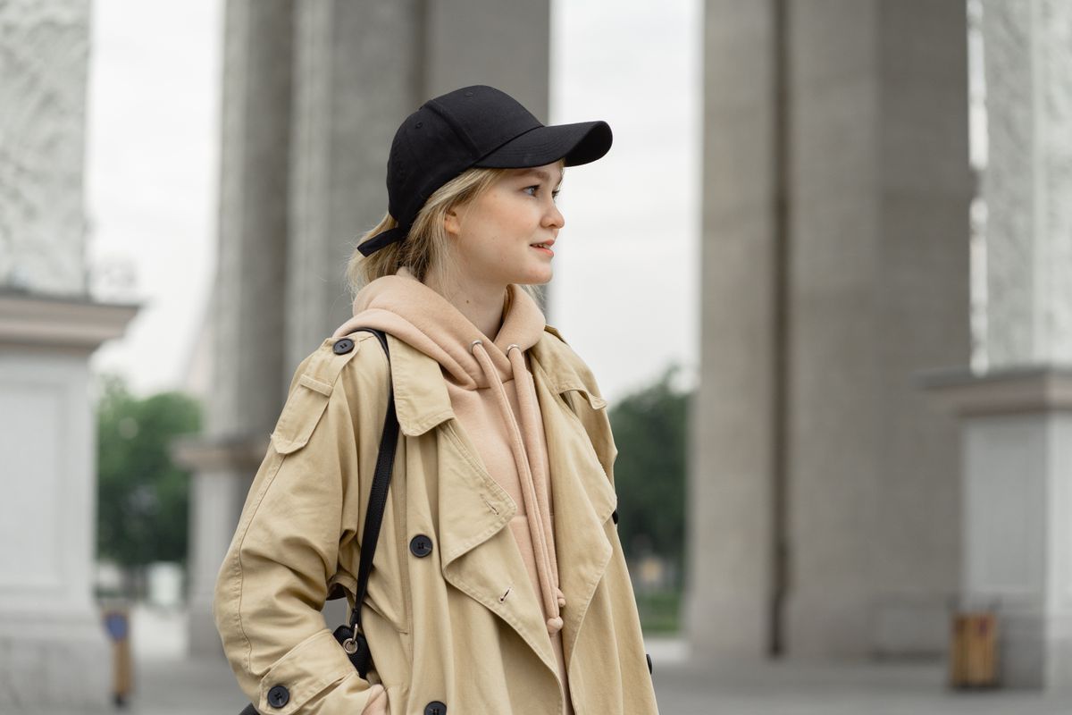 A stylish woman in a trench coat and cap stands outdoors, embodying casual fashion.