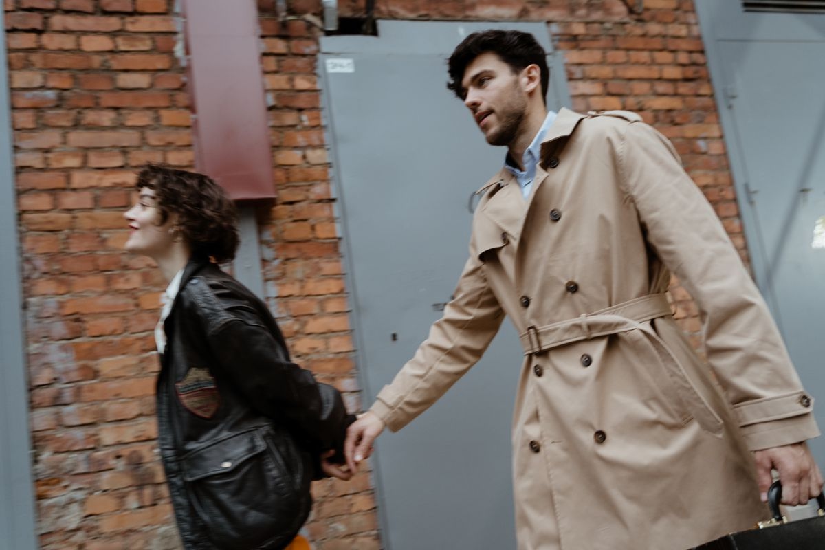 Man and woman walking hand in hand by a brick wall, carrying a suitcase.