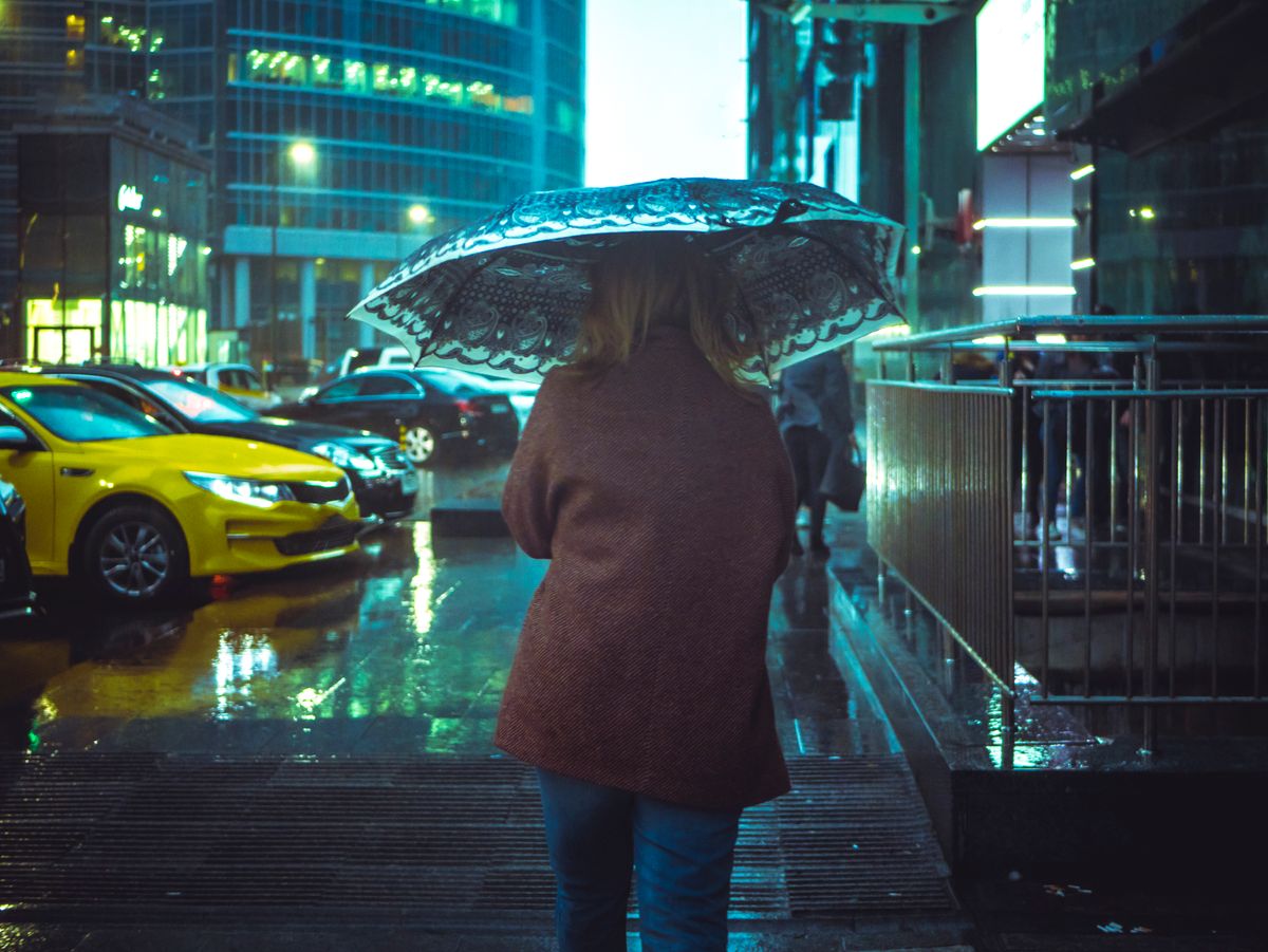 A woman walks under an umbrella on a rainy city street at night, reflecting urban life.