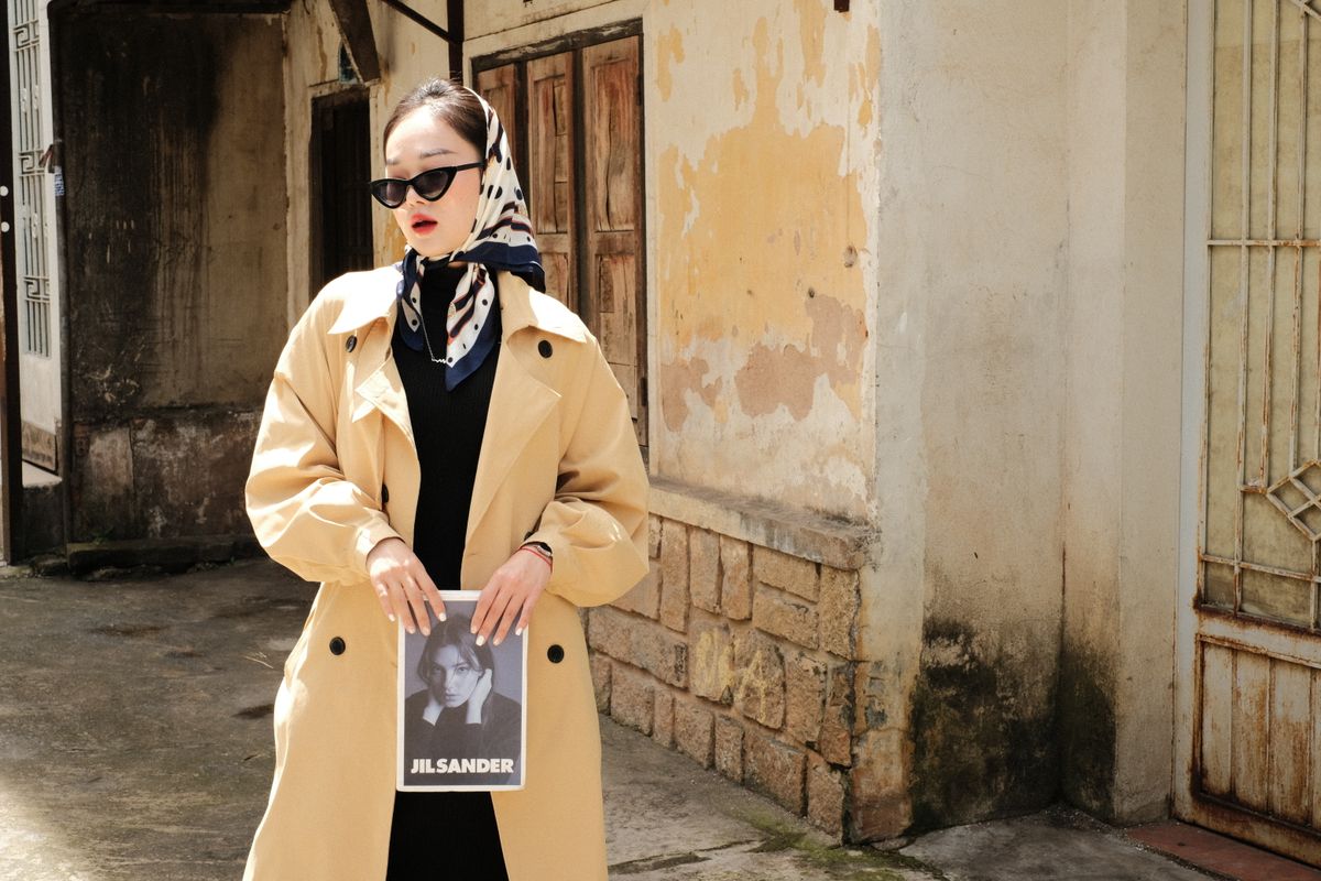 Woman in fashionable trench coat and headscarf holding magazine, posing on a rustic street.