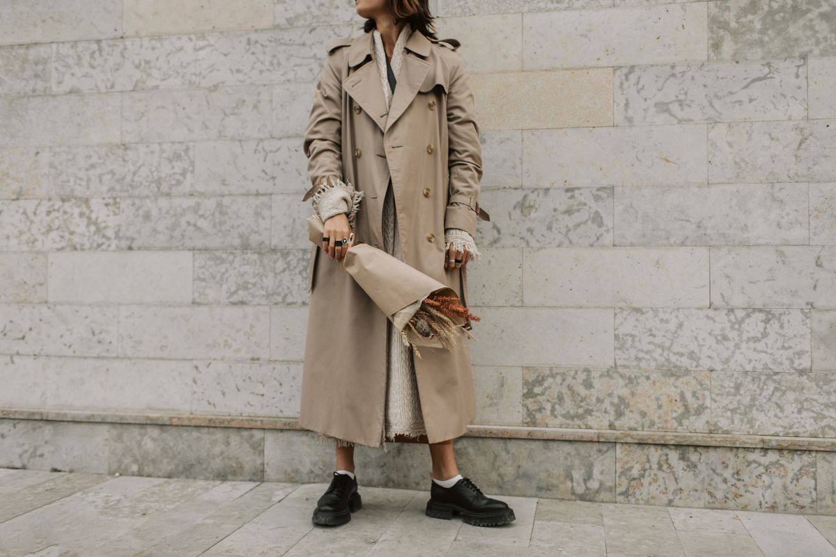 Stylish woman in beige trench coat holding a bouquet against a marble wall. Elegant urban fashion vibe.