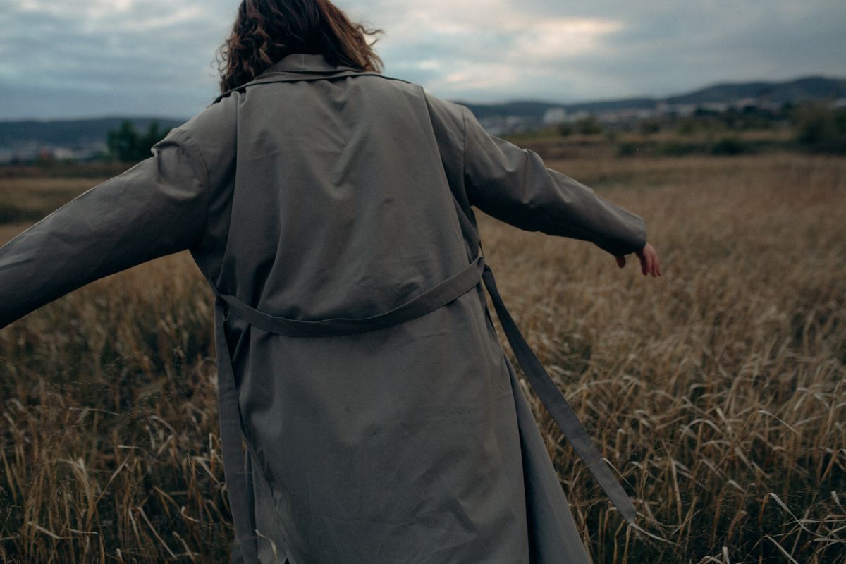 A woman in a long coat walking through a grassy field during a cloudy day.