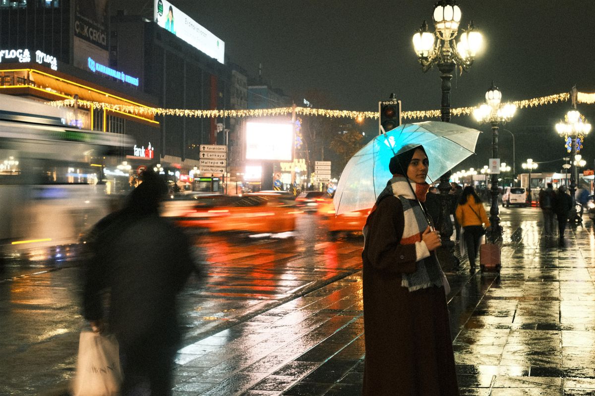 A woman stands with an umbrella at night on a rainy urban street.