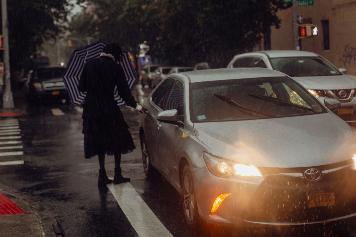 A woman with an umbrella stands near a car during a rainy day in a city.