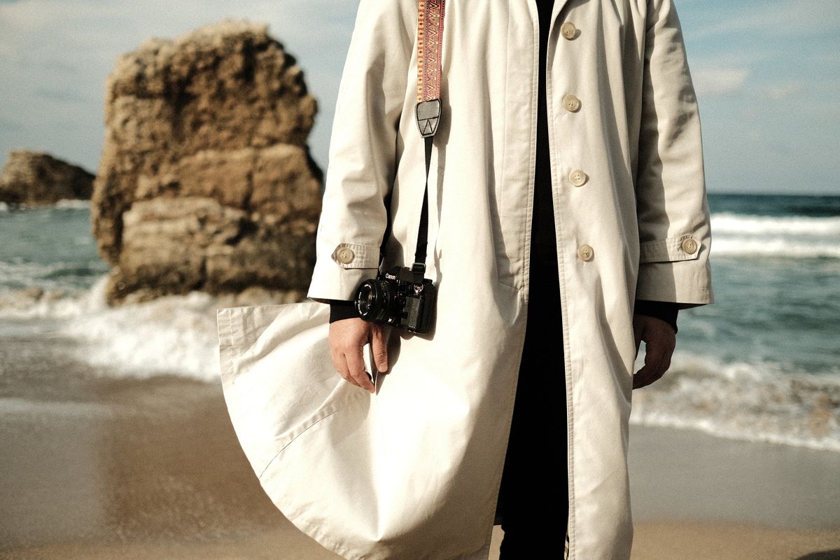A man in a trench coat holding a camera stands by the rocky coast.