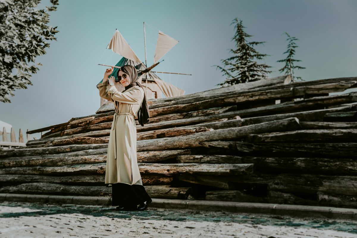 Fashionable woman in trench coat and sunglasses stands near rustic windmill on a sunny day.