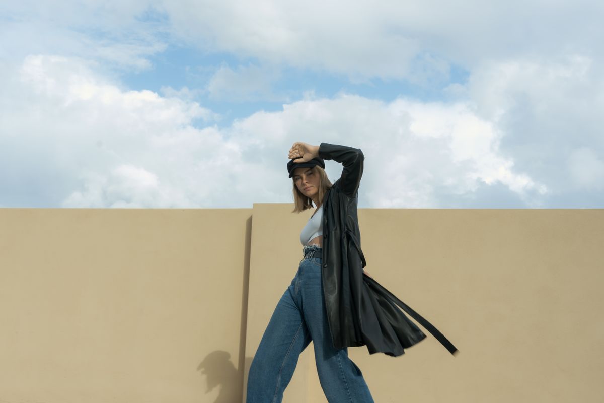 Stylish woman poses against a yellow wall in a leather trench coat and blue jeans under a clear sky.