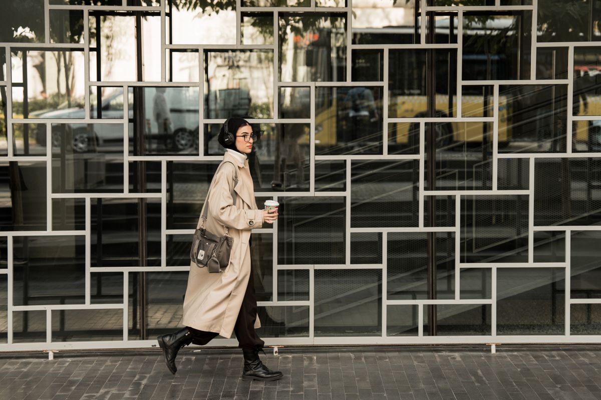 Fashionable woman in beige coat walking with coffee in urban setting. Modern style and urban lifestyle captured outdoors.