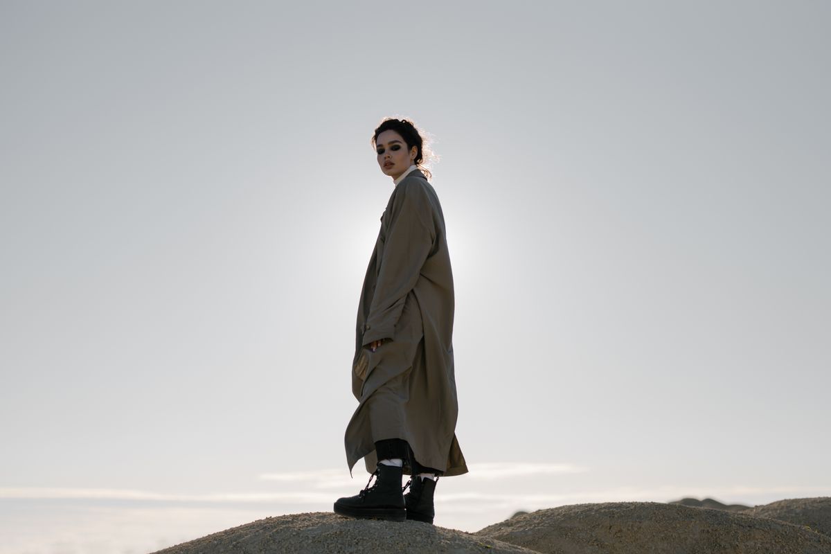 A woman stands on a dune in a trench coat, fashionably silhouetted against the sunlight.