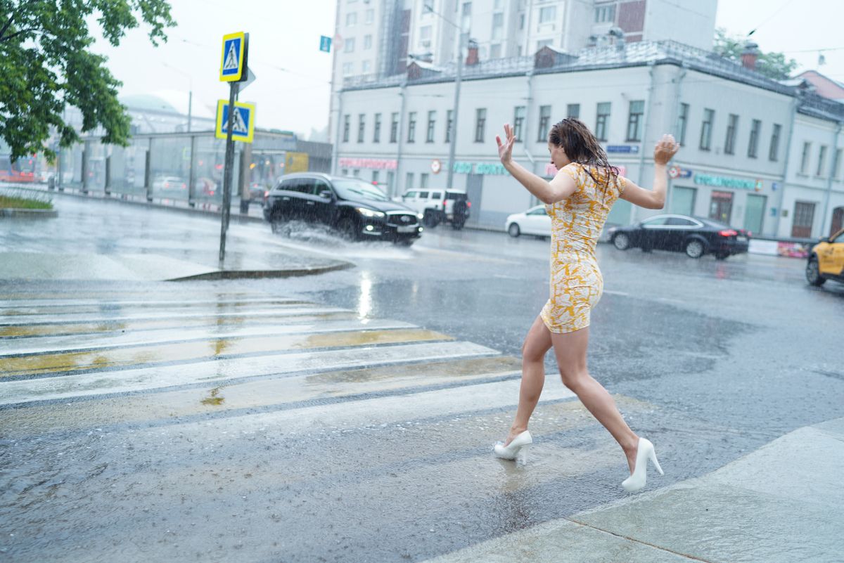 Brunette woman in dress crossing a wet city street in rain, wearing white heels.
