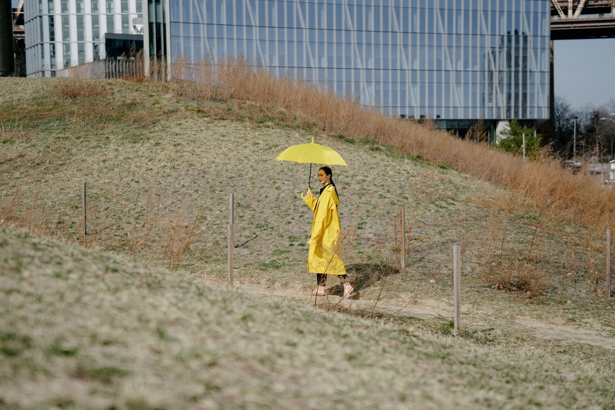 A woman in a yellow coat walks outdoors under a matching umbrella, blending fashion with nature.