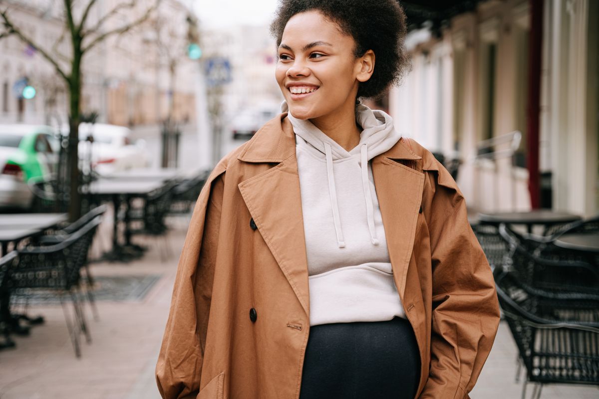 A smiling pregnant woman with afro hair walks on a city sidewalk in a stylish brown coat.