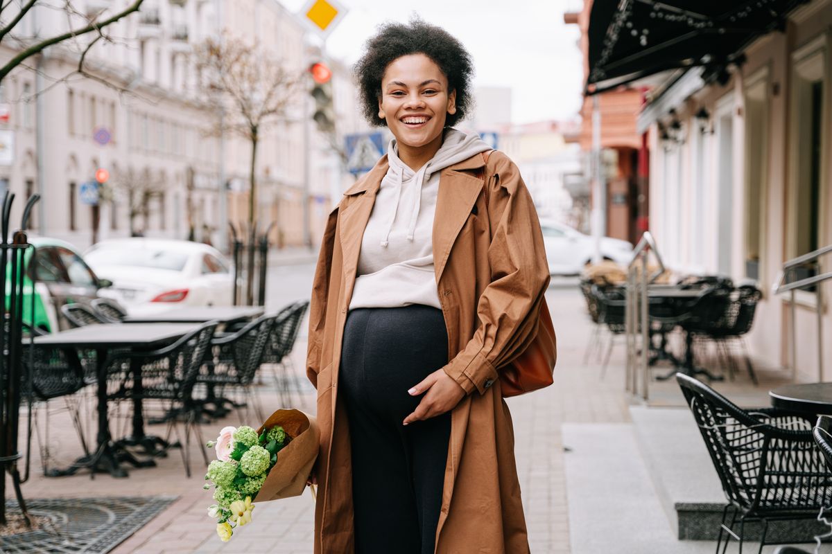A joyful pregnant woman with a bouquet stands on a city sidewalk. Urban maternity fashion.