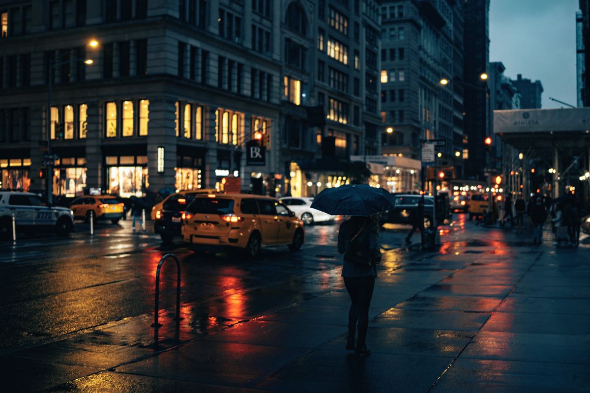A lone woman walks under an umbrella on a rainy city street, surrounded by urban lights.