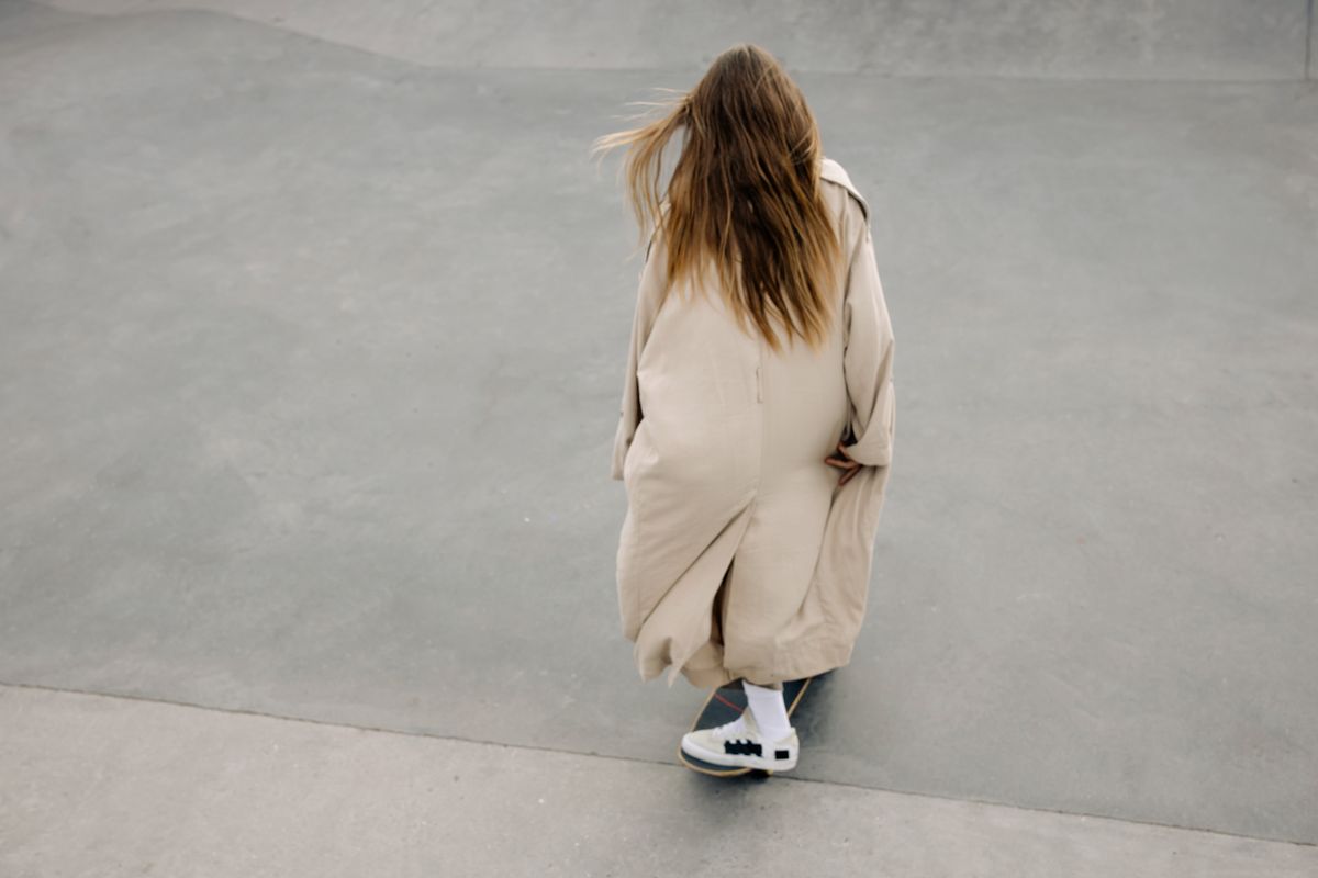 Back view of a woman skateboarding in a trench coat, showcasing motion and style.