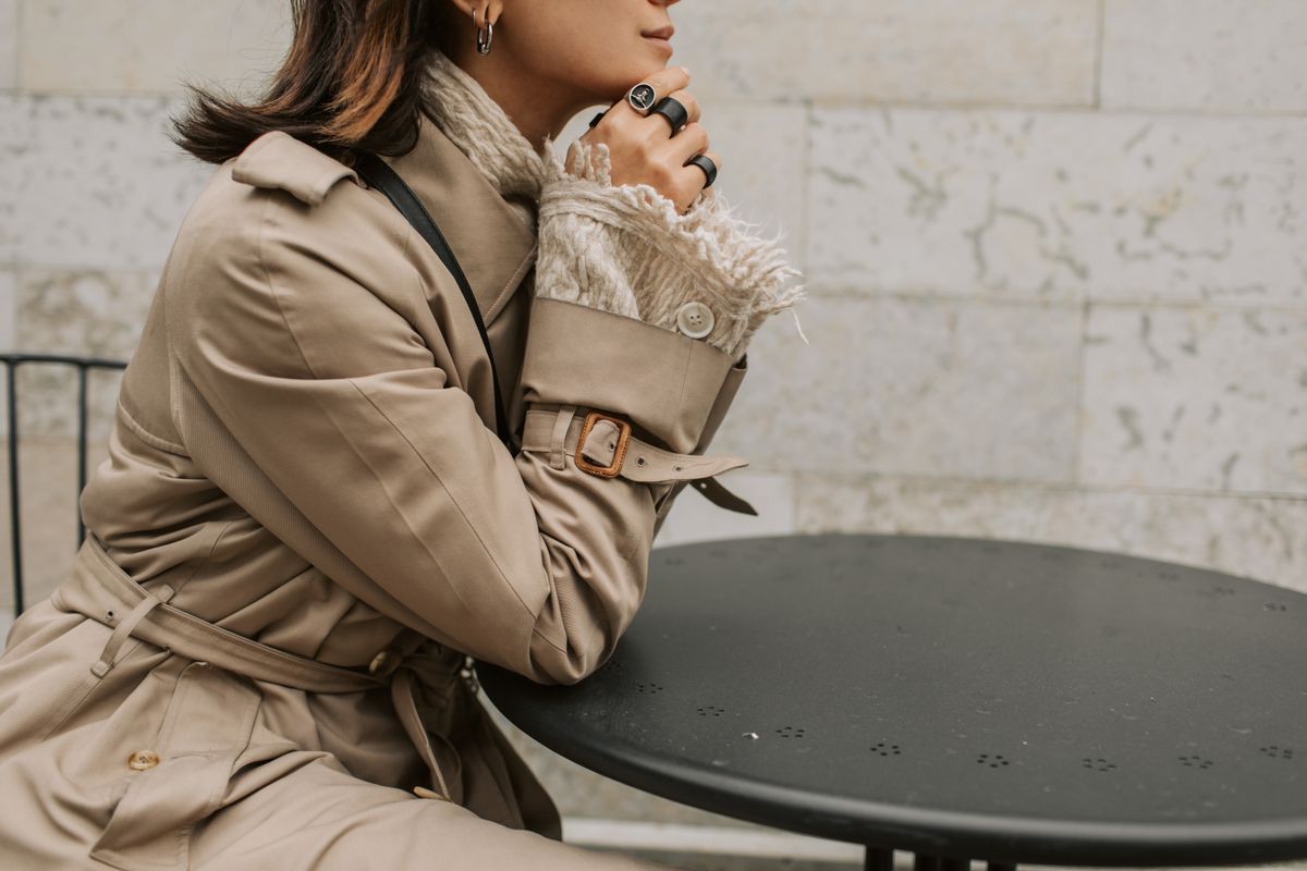A fashionable woman in a beige trench coat sits outdoors at a table during autumn.