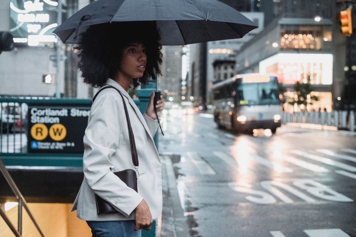 A woman with an umbrella stands on a rainy city street near a subway entrance.
