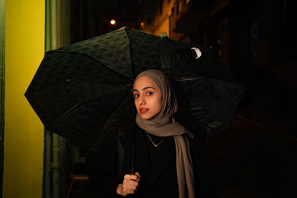 Portrait of a woman holding an umbrella at night in the city, wearing a hijab and standing under a streetlight.