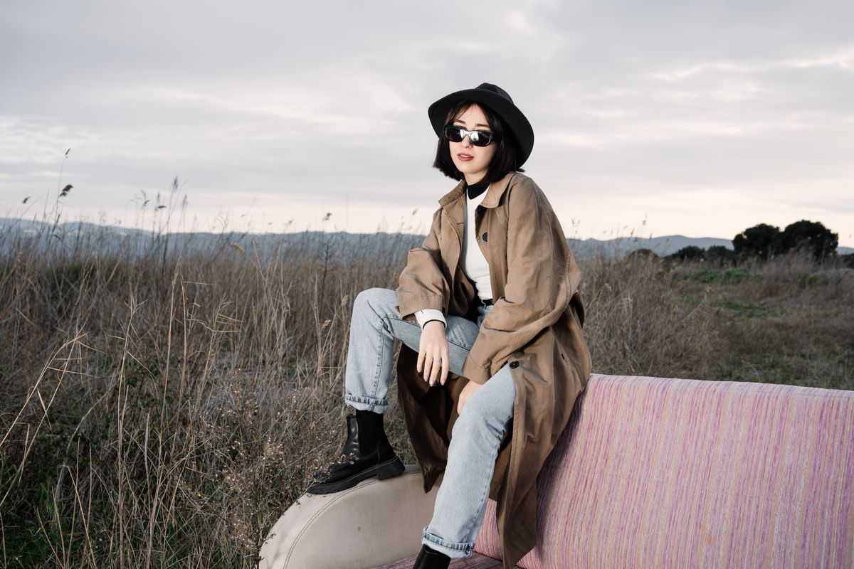 Young woman in trench coat and sunglasses sitting on a couch in a field, showcasing modern fashion style.
