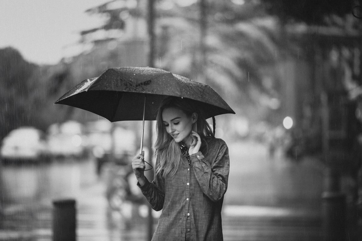 A woman holding an umbrella walks alone on a rainy street, captured in black and white.