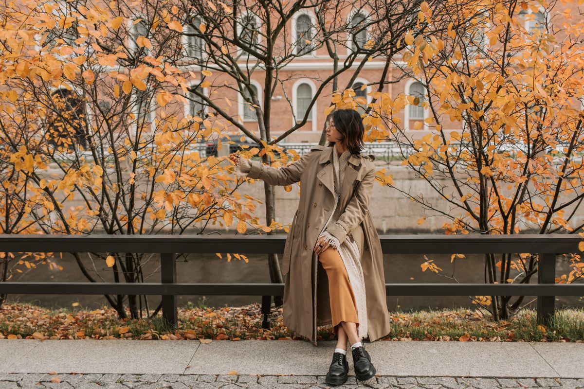 Asian woman enjoying fall foliage while sitting on a bench wearing a trench coat.