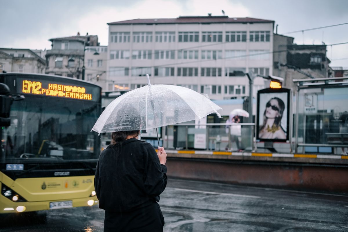 A woman stands with an umbrella at an İstanbul bus stop on a rainy day.