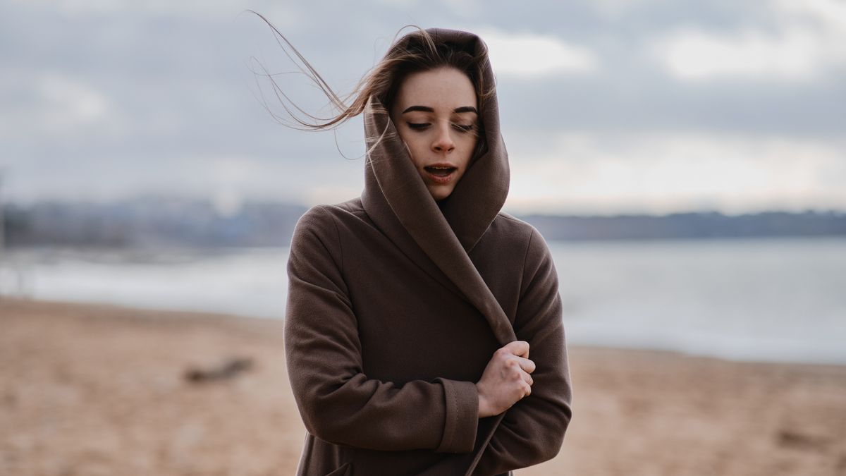 A woman stands on a windy beach with her coat wrapped around her, capturing a serene winter moment.