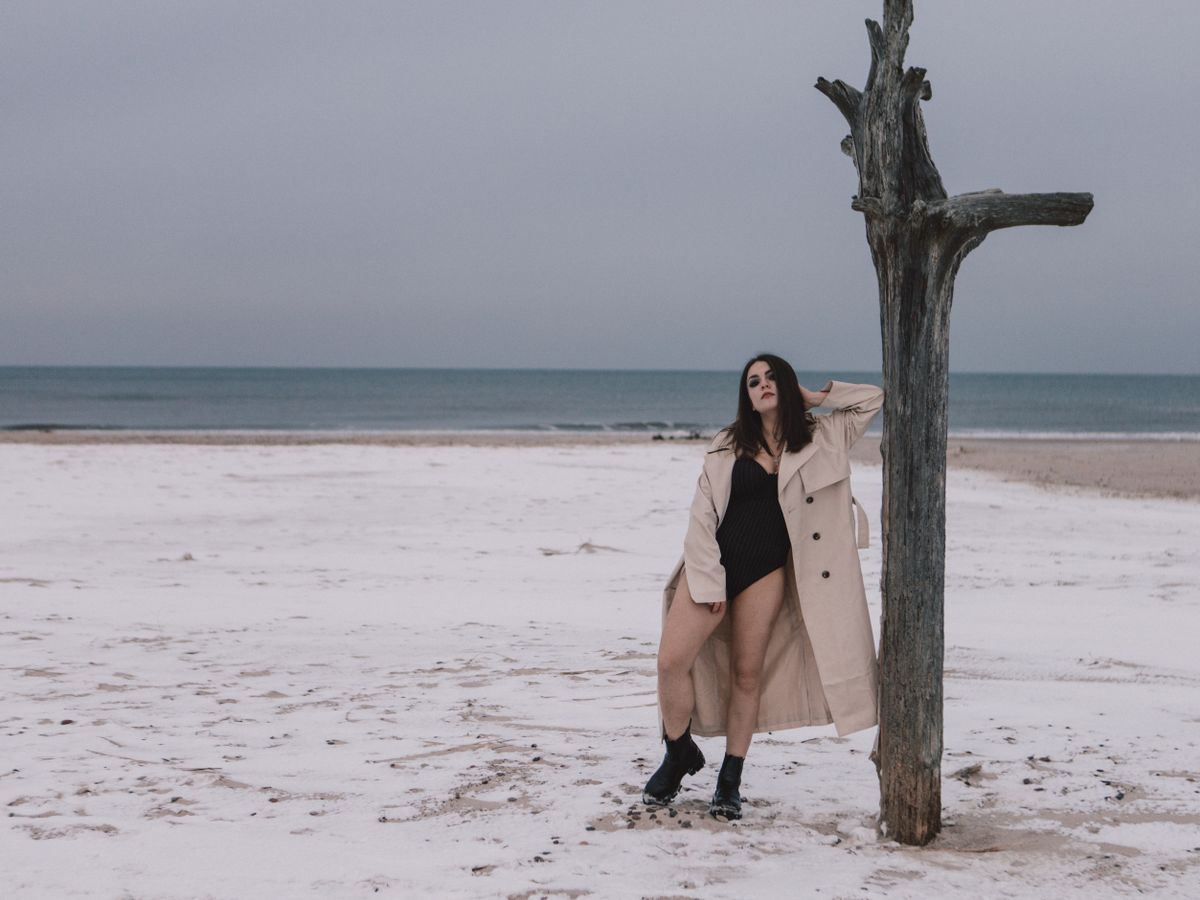 Model in trench coat poses on a snowy beach with withered tree during winter.
