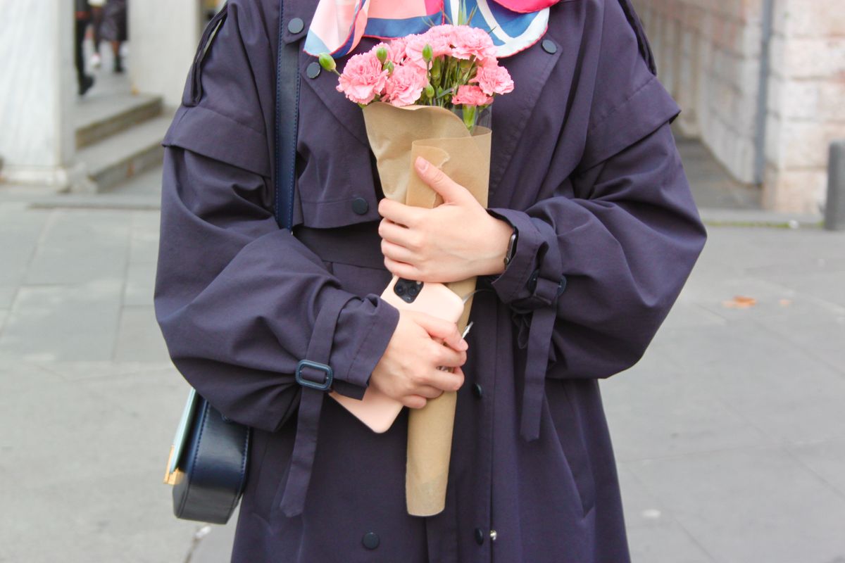 A woman in a dark coat holds a bouquet of pink flowers outdoors on a city street.