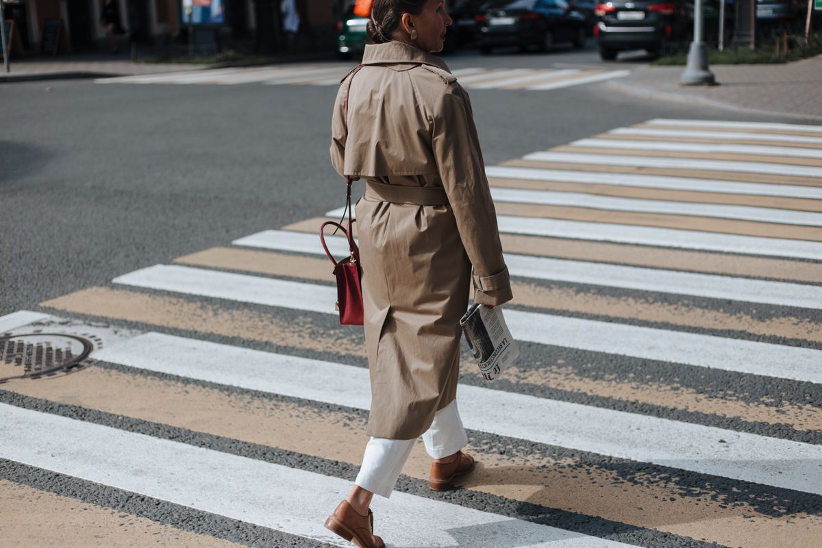 A stylish woman crosses a city street in a trench coat, holding a newspaper.