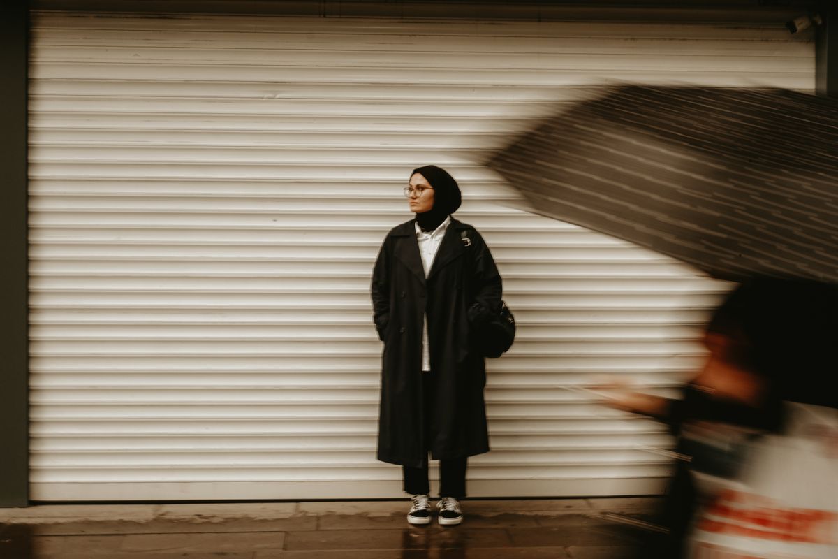A woman in a black coat stands in a city street against a shuttered wall as blurred motion passes by.
