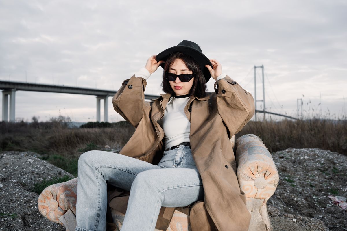 A woman in sunglasses and trench coat posing outdoors against a bridge backdrop.