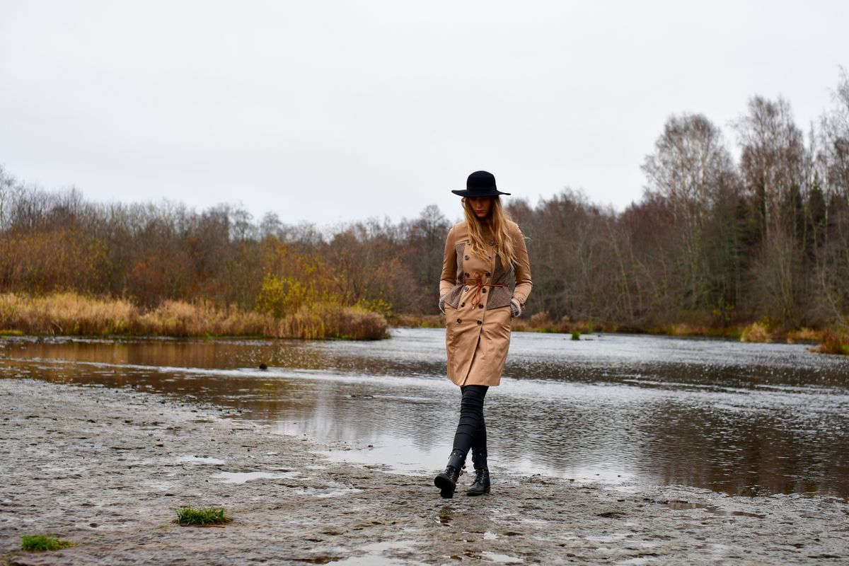 A woman in a trench coat and hat walks along the Jägala River in Estonia during fall, surrounded by autumn foliage.