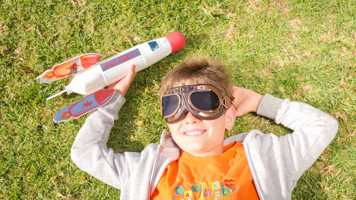A young boy lying on grass, wearing goggles, and playing with a toy rocket, smiling in the sun.