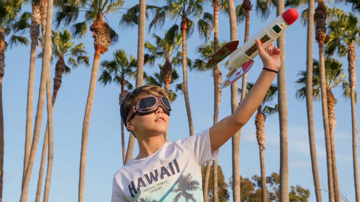 A child joyfully playing with a toy rocket under palm trees on a sunny day.