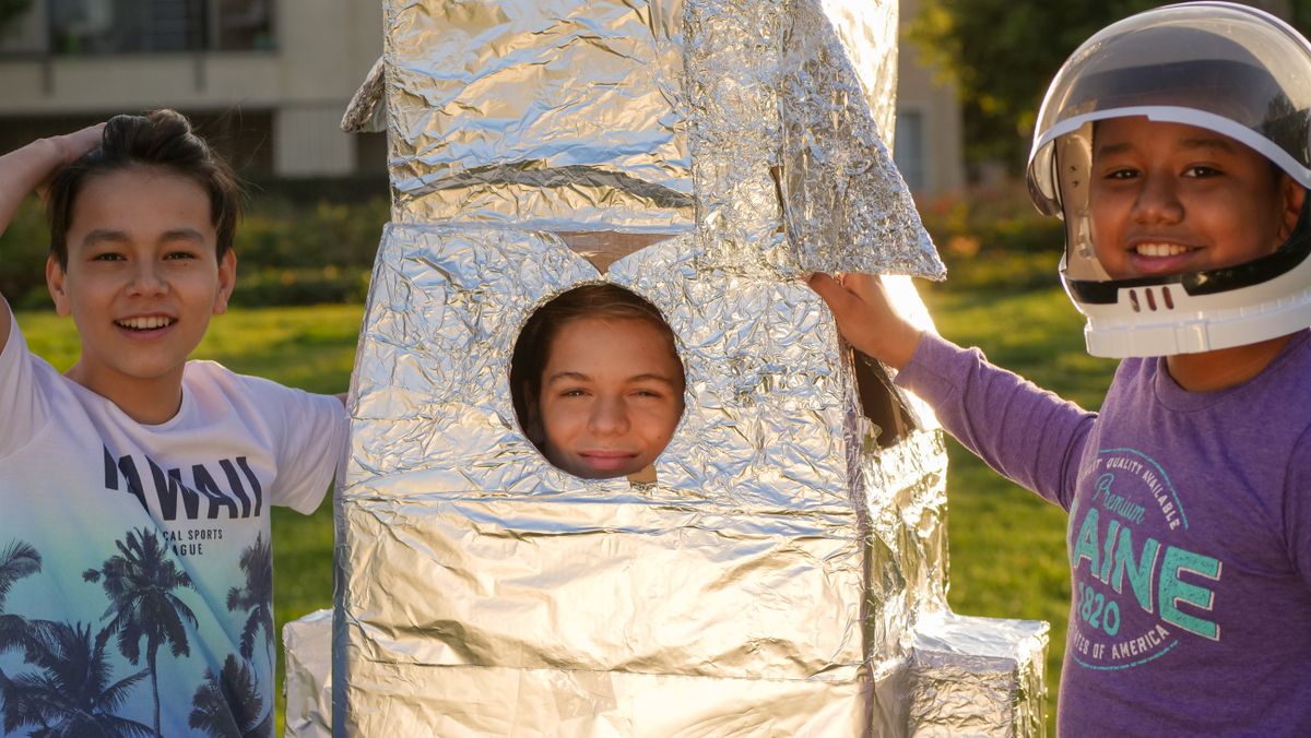 Three kids having fun with a cardboard rocket outdoors during the day.