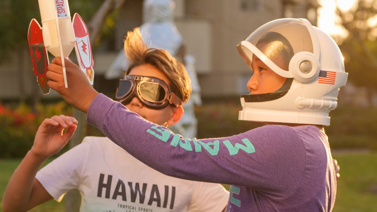 Two kids engage in imaginative play with astronaut gear and rocket toy outdoors.