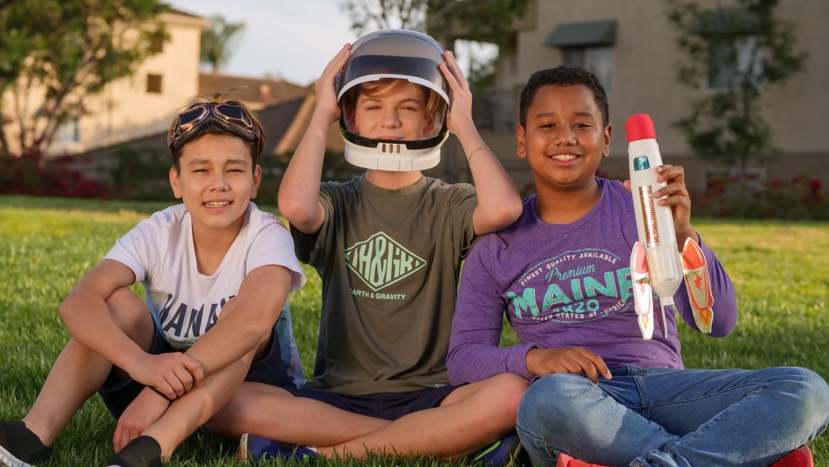 Three kids sitting on grass, playing with a toy rocket, enjoying a sunny day.